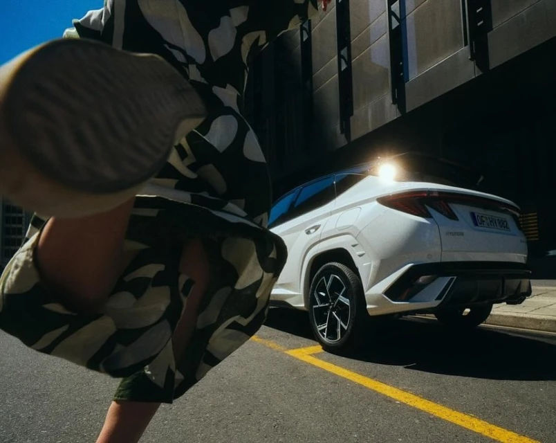 White SUV car photographed from the rear on asphalt road with blurred woman's legs. Building in the background and golden light.
