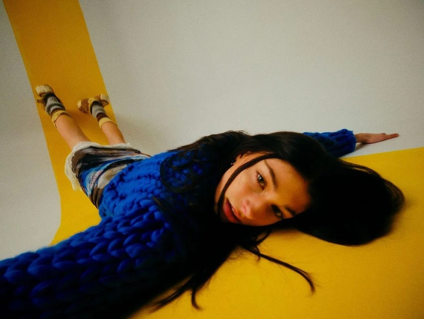 Young woman in a chunky blue knit sweater lies on a yellow surface with white walls, legs raised, posing with outstretched arms.