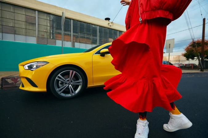 Person wearing a bright red dress and white sneakers walking past a parked yellow sedan on a city street.