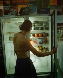 Woman in red halter top and blue skirt looking inside an open convenience store refrigerator with packaged goods