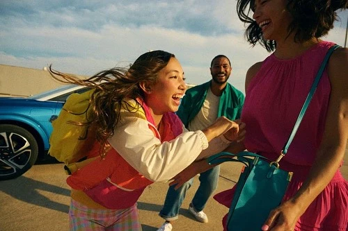 Four people laughing and playing outdoors near a blue car with bright sunlight casting warm tones.