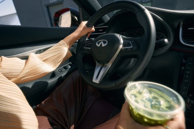 Woman driving an Infiniti car holding a green drink in a plastic cup, close-up view of the steering wheel and car interior.