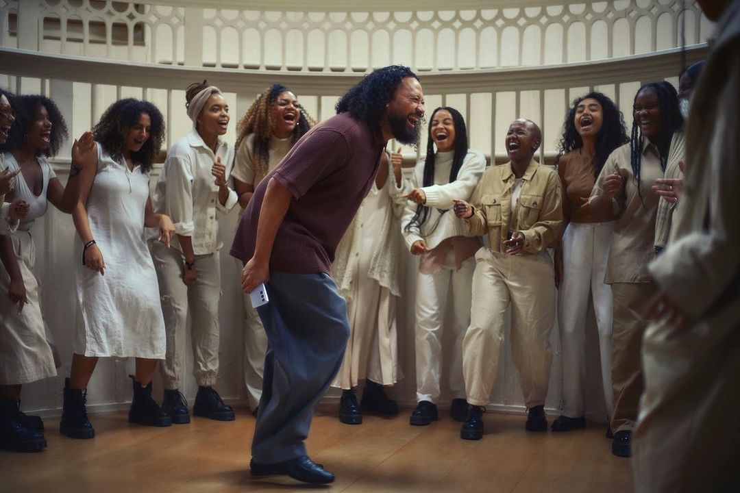 Enthusiastic man leads a group of women in song inside a building with arched windows; smiles and joyful expressions fill the scene.