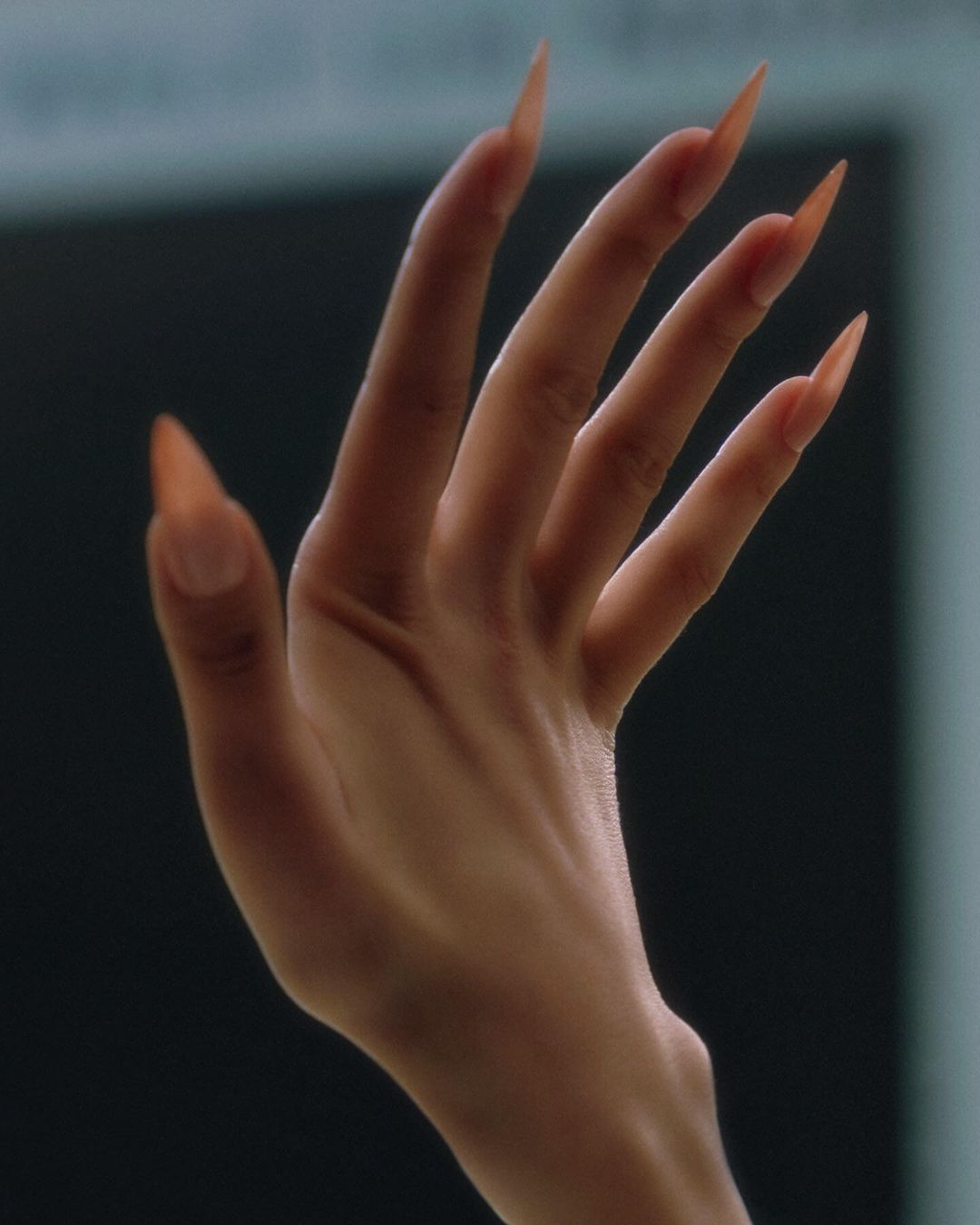 Close-up of a tan adult female hand with long, pointed orange fingernails is displayed against a blurred, dark background.