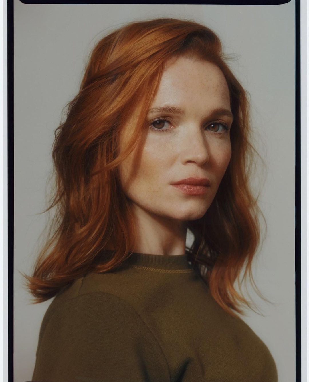 Portrait of a fair-skinned adult female with red hair, wearing an olive green t-shirt, posing in front of a neutral background.