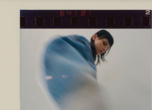 Woman with dark hair stands in a blurred blue and white scene; film strip along the top edge visible in a medium-shot composition.