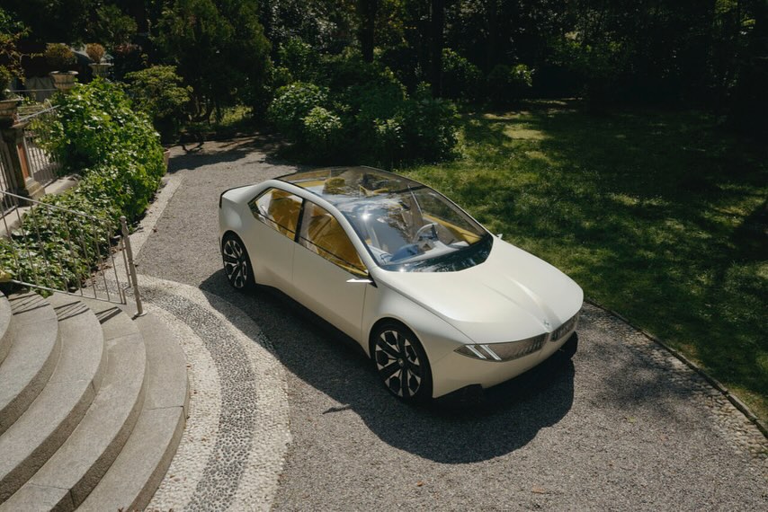 Exterior of a modern, white BMW concept car with a transparent roof parked on a gravel driveway surrounded by lush greenery.