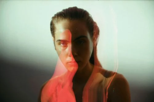 Close-up portrait of a woman with fair skin and dark hair, red and white lighting, against a pale, neutral background in a studio.