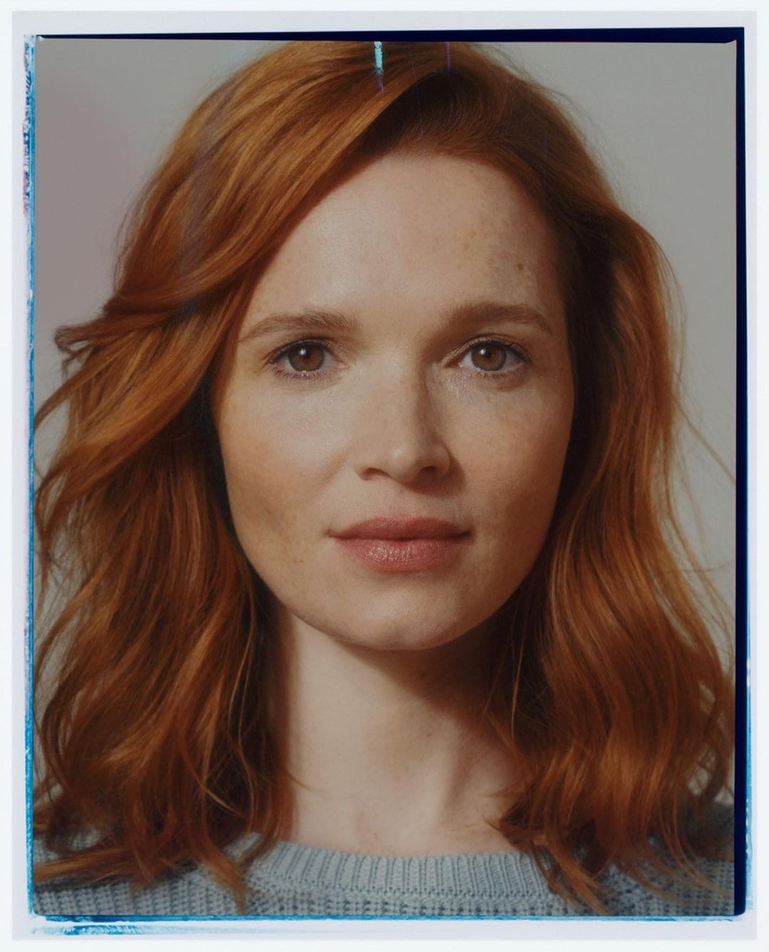 Close-up portrait of a red-haired woman with freckles, gazing directly at the camera wearing a grey top against a neutral background.