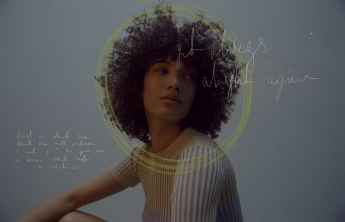 Portrait of a black woman with an afro, wearing a gray top, posing in a photo studio against a dark gray background.
