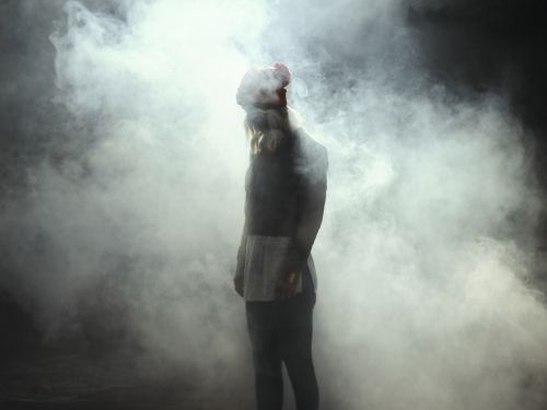 Full-length shot of a person in dark clothing and a red hat standing in a smoky haze against a dark background. 
