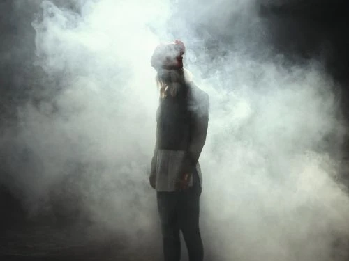 Full-length shot of a person in dark clothing and a red hat standing in a smoky haze against a dark background. 