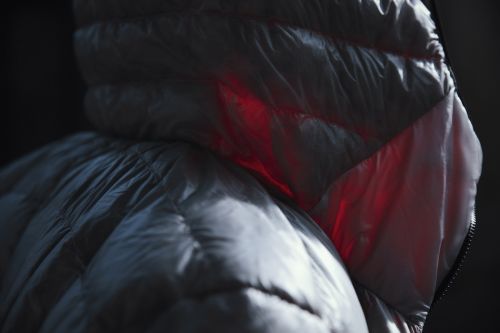 Close-up of a puffy, light gray jacket with a high collar illuminated by a red light, against a dark background. Fabric texture is detailed.