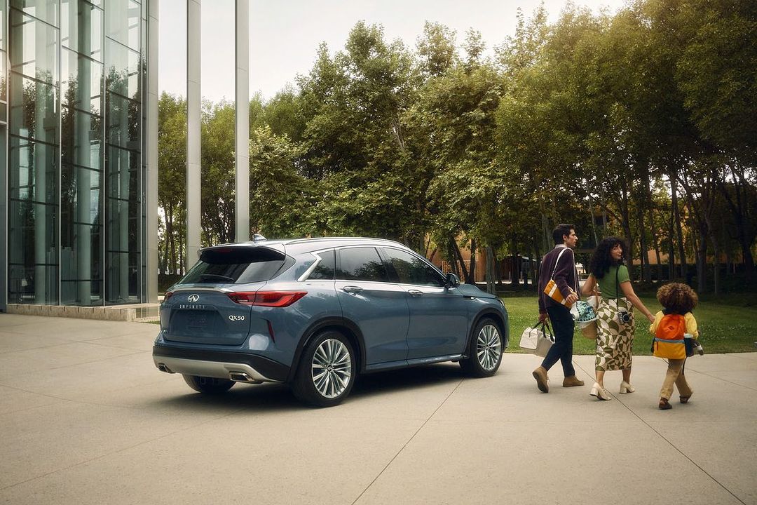 Blue Infiniti QX30 parked outside a glass building as a family walks by on a sunny day. Automotive lifestyle imagery.