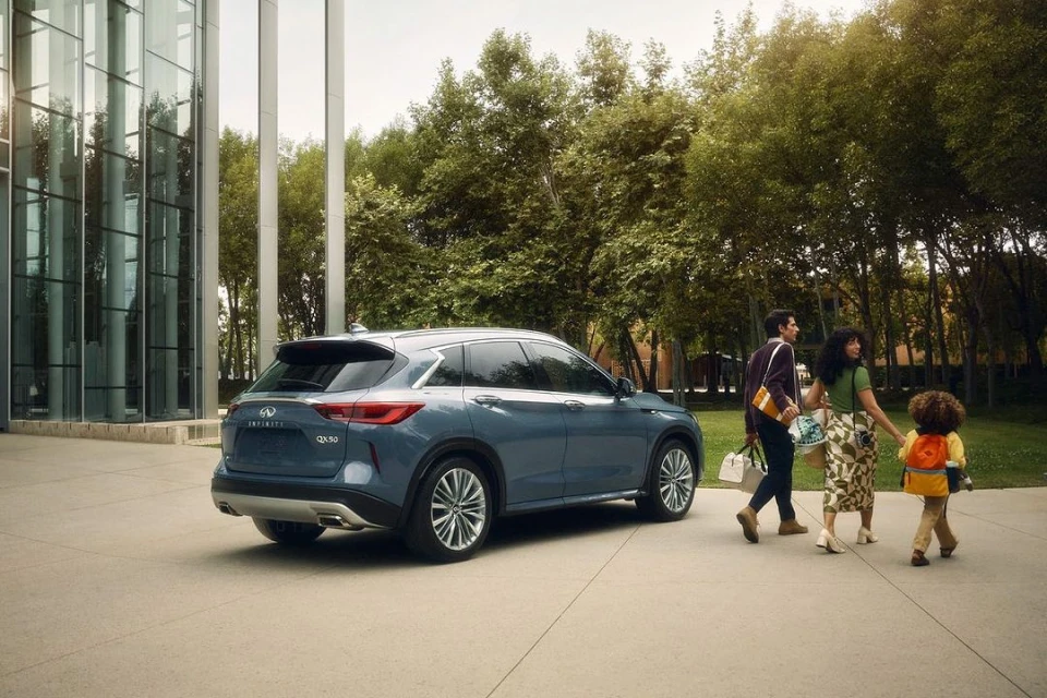 Blue Infiniti QX30 parked outside a glass building as a family walks by on a sunny day. Automotive lifestyle imagery.