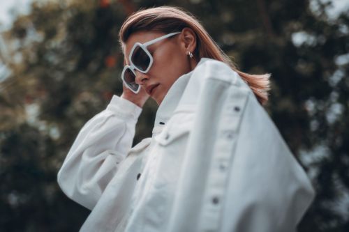 Stylish adult woman in white jacket and angular sunglasses poses outdoors with blurred green tree background. Fashion editorial shot.