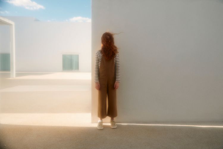 A young, red-haired person in brown overalls stands beside a white wall in a minimalist outdoor setting with bright, diffused light.