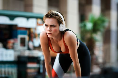 Woman in workout clothes rests with hands on knees outside; she wears headphones and an orange top with black leggings.
