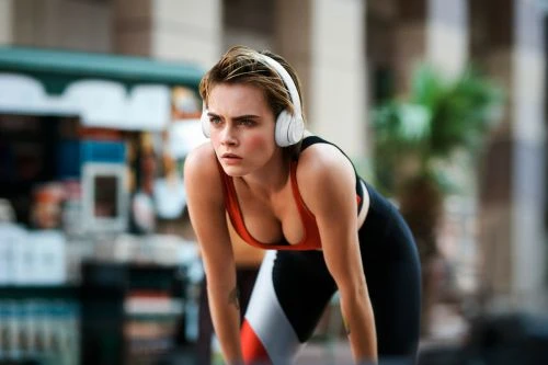 Woman in workout clothes rests with hands on knees outside; she wears headphones and an orange top with black leggings.