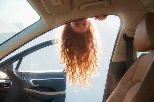Upside down view of a person with red hair inside of a car with an open door, car interior, and bright sunlight in the background.