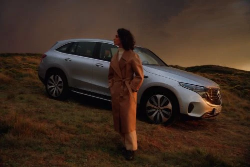 Silver SUV with woman in tan coat on grassy hillside against a dark sky, highlighting automotive design and outdoor lifestyle.