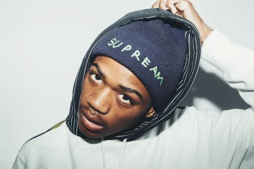 Close-up of African American man wearing blue Supreme beanie and white hoodie, looking at the camera against a white background.
