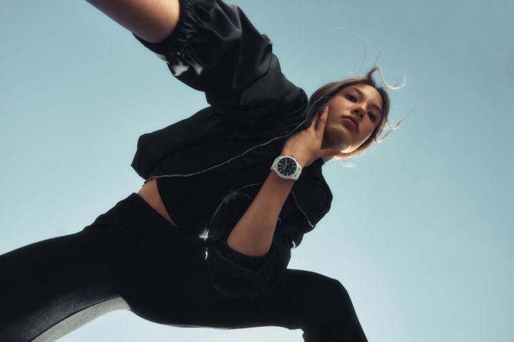 Woman in black crop top and leggings poses against bright blue sky, showcasing a white wristwatch in a low-angle outdoor fashion shot.