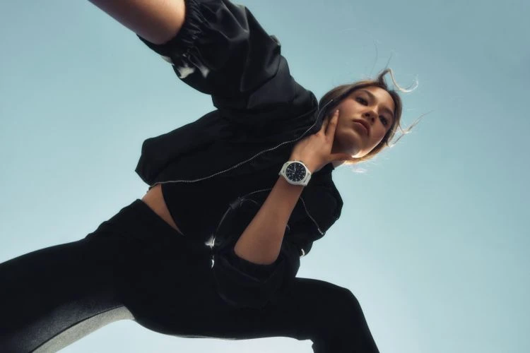 Woman in black crop top and leggings poses against bright blue sky, showcasing a white wristwatch in a low-angle outdoor fashion shot.
