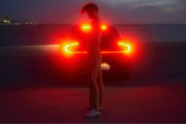 Adult stands near car on beach as taillights glow at night. The scene blends a relaxed coastal setting with the allure of automotive design.
