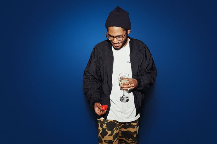 Adult male in a beanie holds a wine glass and strawberry, standing against a royal blue backdrop indoors. He wears glasses and a jacket.