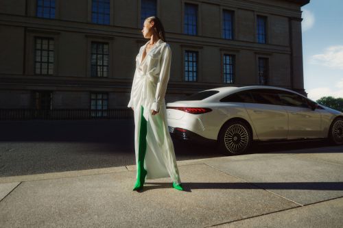 Model in a white dress and green boots standing on concrete next to a parked white car and classical building