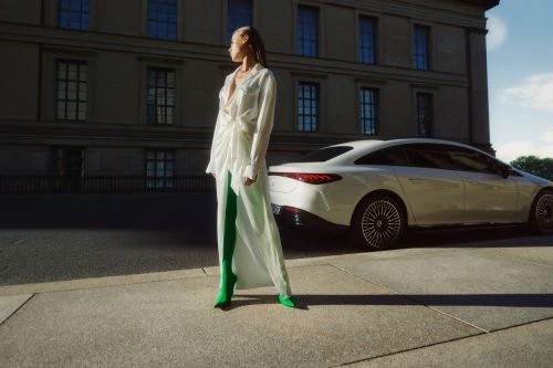 Model in a white dress and green boots standing on concrete next to a parked white car and classical building