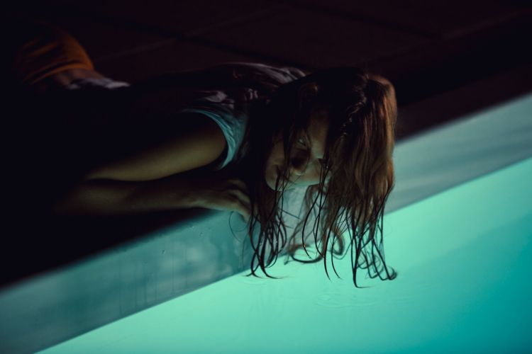 An adult female, appearing wet, is positioned at the edge of a bright turquoise-colored pool, under low lighting, creating a dramatic scene.