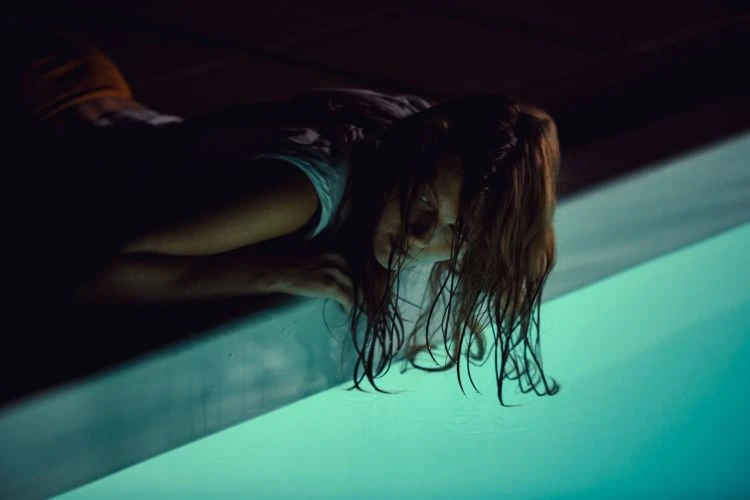 An adult female, appearing wet, is positioned at the edge of a bright turquoise-colored pool, under low lighting, creating a dramatic scene.