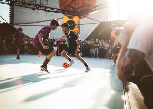 Two soccer players compete for the ball during a game on an indoor court in front of a crowd of spectators and orange balloons.