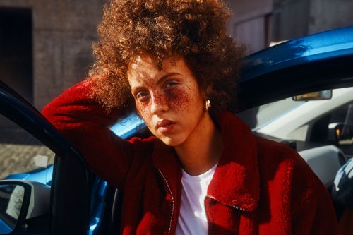 Young woman with curly red hair in red coat poses with blue car outdoors in sunlight. Beauty and fashion visual content.