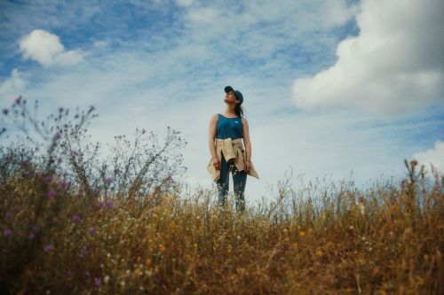 Woman in a blue tank top and baseball cap standing outdoors in a grassy field looking up against a cloudy sky