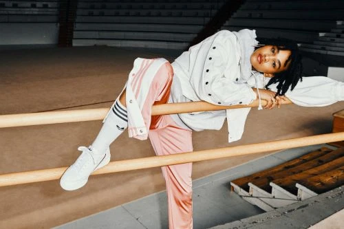 A woman with dreadlocks wearing Adidas athletic apparel poses across a wooden railing in an indoor arena with concrete and grey bleacher seating.