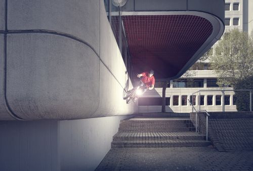 BMX rider performs mid-air trick on bike against urban concrete architecture with stairs and distant buildings in an outdoor setting.