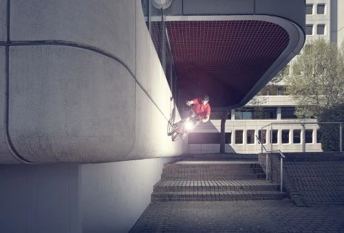 BMX rider performs mid-air trick on bike against urban concrete architecture with stairs and distant buildings in an outdoor setting.