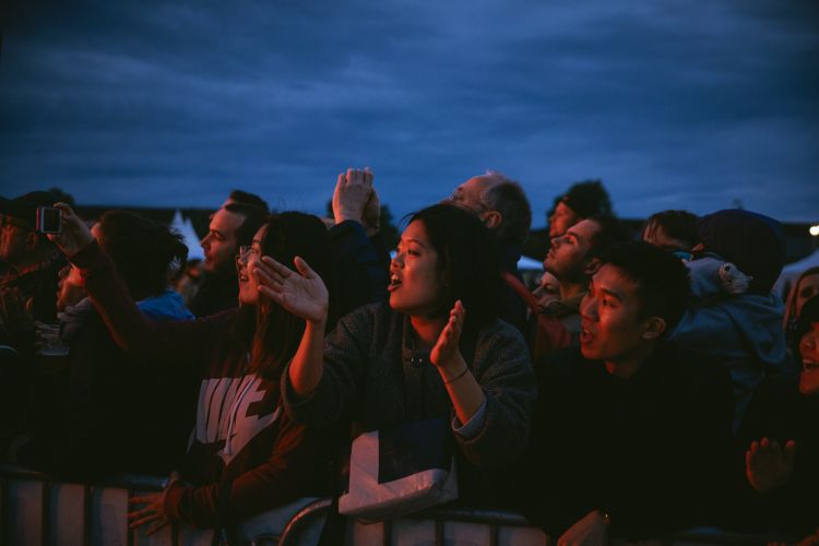 Enthusiastic crowd of diverse adults celebrates at an outdoor event with visible fencing in the foreground and cloudy sky at dusk.
