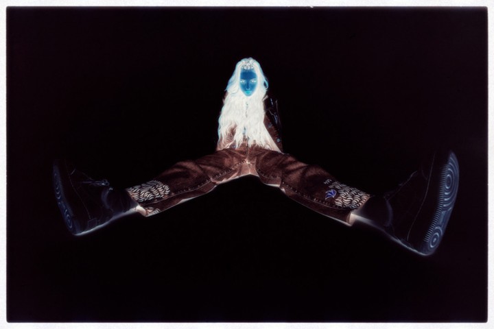 Inverted portrait of a person with long white hair wearing brown pants and black sneakers against a black background.