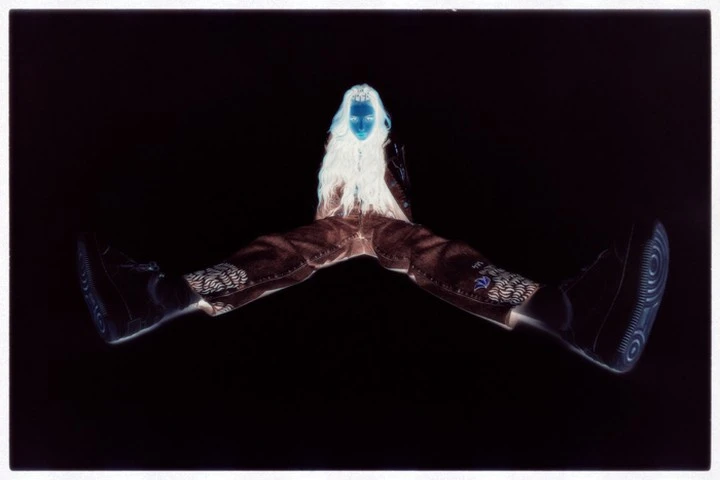 Inverted portrait of a person with long white hair wearing brown pants and black sneakers against a black background.
