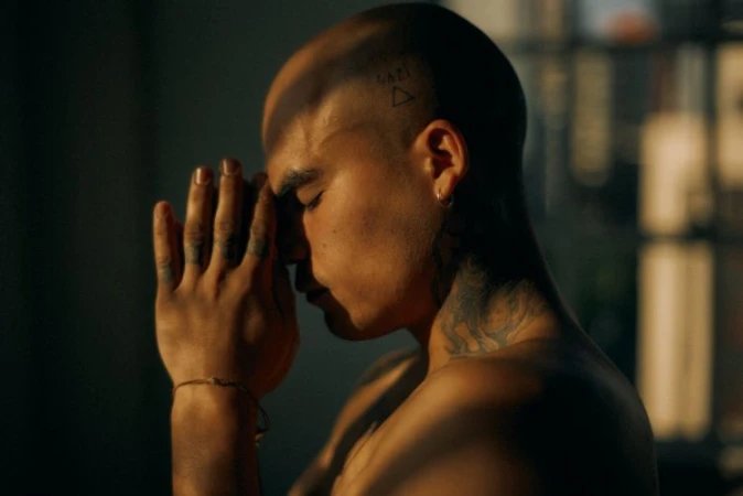 Close-up of a bald adult male in a dimly lit indoor space, wearing earrings, neck tattoos, and a bracelet, with hands clasped in prayer.