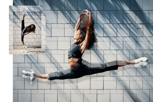 A woman in a black outfit does the splits against a textured white tile wall, bathed in natural light with shadows, capturing movement.
