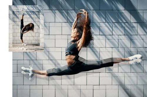A woman in a black outfit does the splits against a textured white tile wall, bathed in natural light with shadows, capturing movement.