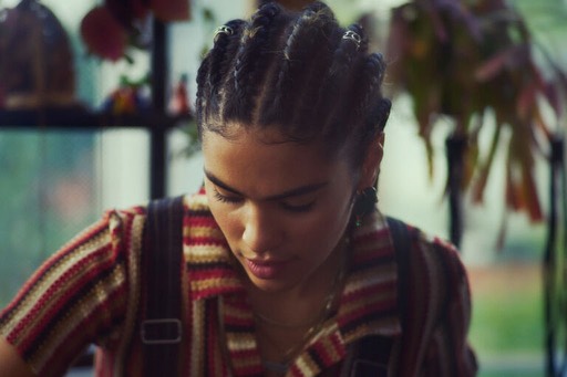 Close-up of a young woman with braided hair, wearing a striped shirt and overalls, looking down in a bright indoor setting.