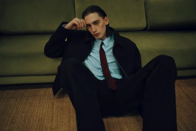 Young man with dark hair seated on a textured floor leaning against a green sofa, wearing a black jacket, light blue shirt, and dark tie.