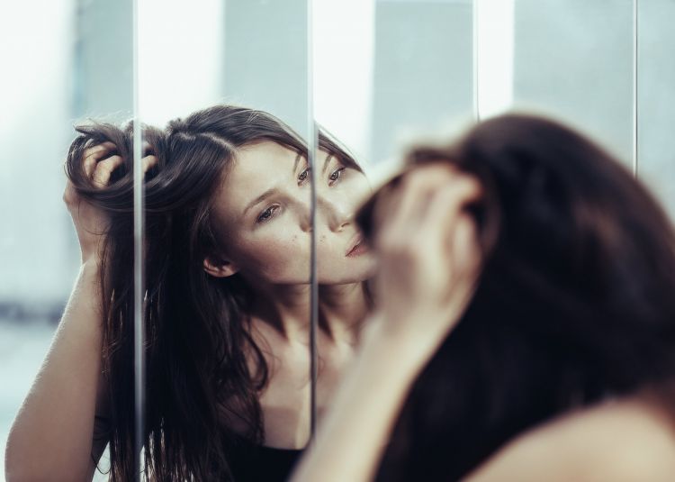 Woman gazes into a paneled mirror, her hand touching her dark hair. Reflections create layered perspective in a naturally lit, indoor setting.