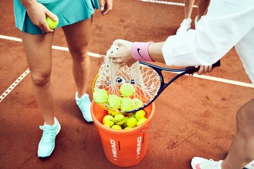 Two women on a tennis court, one holding a bucket filled with tennis balls and the other a racket; athletic wear and tennis shoes are visible.